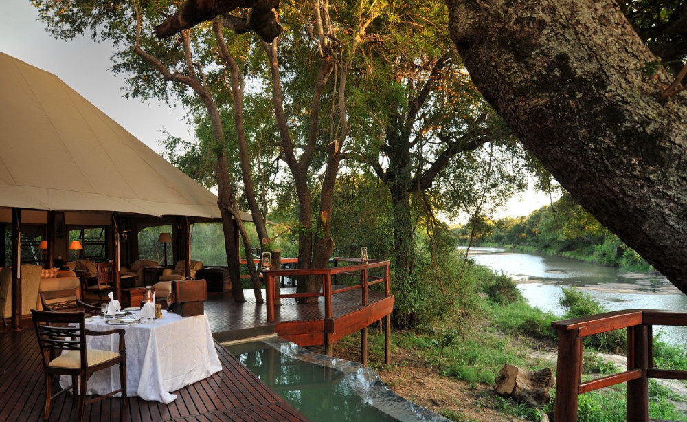 A luxurious tented camp deck with a dining table, surrounded by trees and overlooking a river, captured during a BST Tours safari in Mluwati Concession.