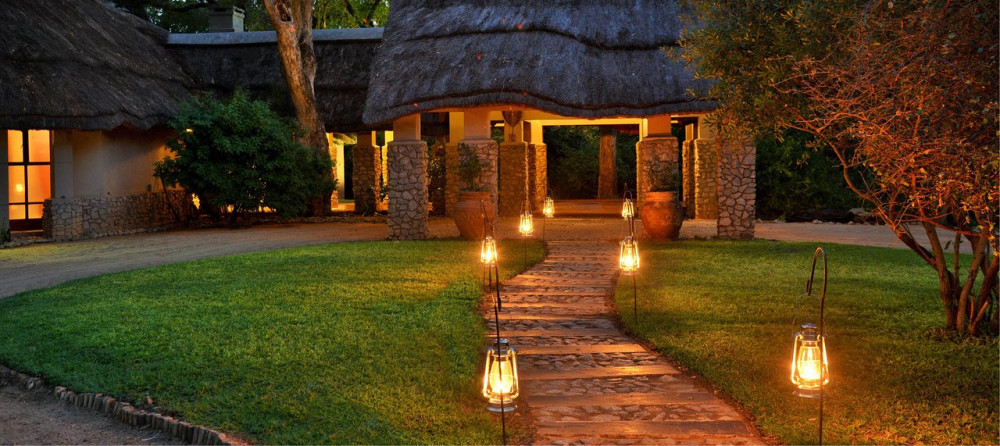 A lantern-lit stone pathway leading to a thatched-roof lodge surrounded by lush grass and trees, captured at Imbali Safari Lodge during a BST Tours safari in Kruger National Park.